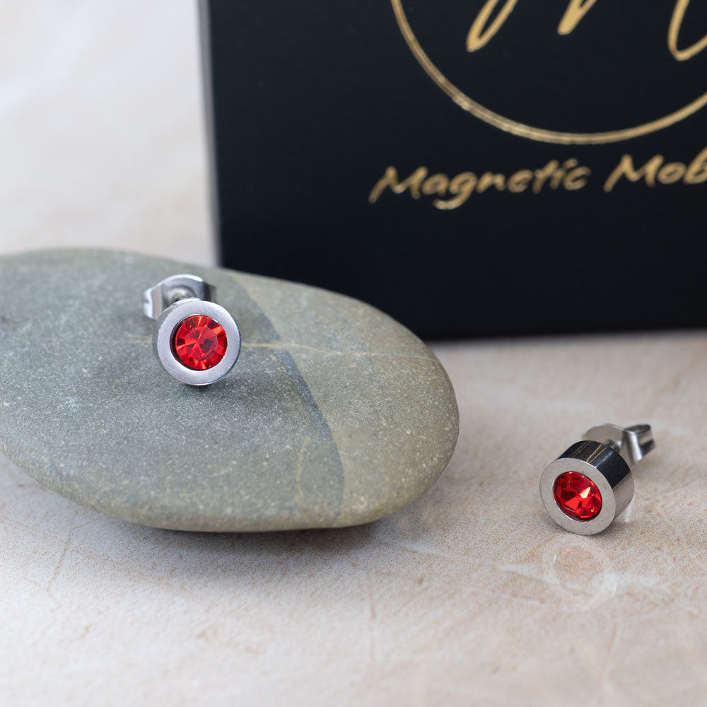 Magnetic Mobility Birthstone Stud Earrings featuring red Swarovski crystals. These earrings have a magnetic back and are ideal for providing stylish pain relief. Displayed on a smooth stone with Magnetic Mobility branded box in the background.